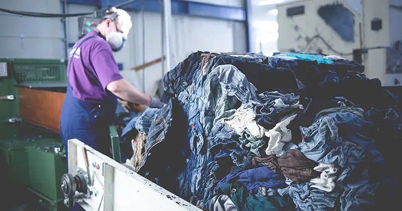 Een werknemer met een mondkapje sorteert in een fabrieksomgeving een grote stapel gebruikte kleding, waarschijnlijk met het oog op recycling of textielverwerking.